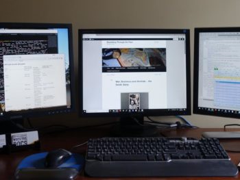 3 monitors on a desk with a mug, mouse, keyboard and open book with glasses on it.